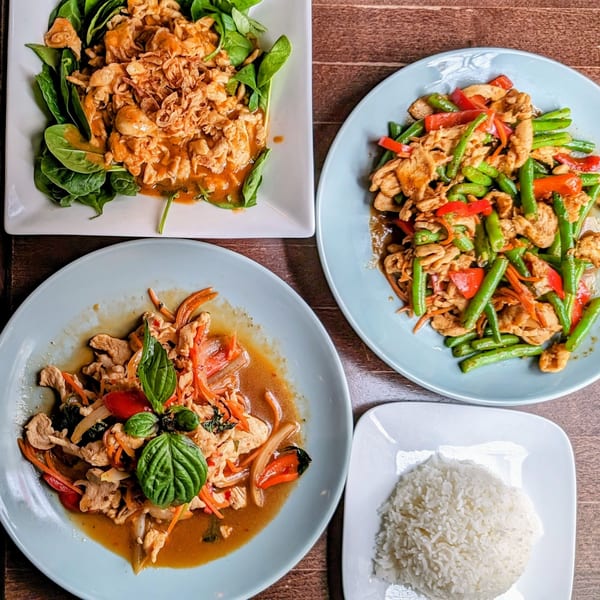 Three plates of Thai chicken dishes with vegetables and a side of white rice on a wooden table.